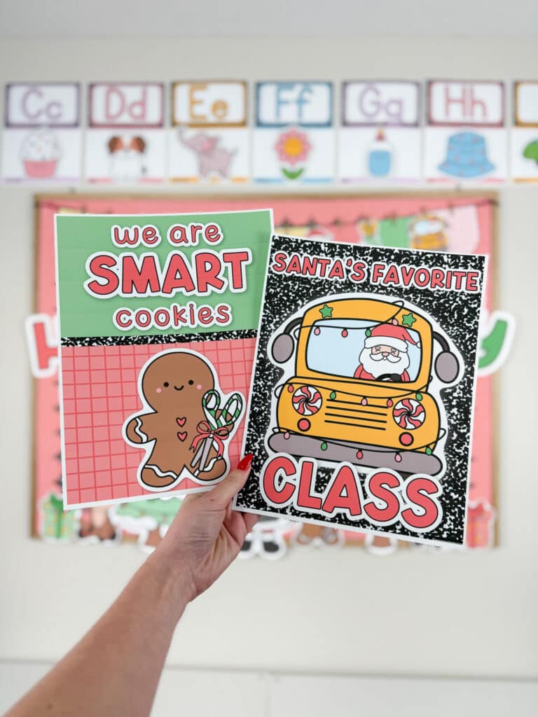 A hand holds two festive Christmas classroom decor posters. One poster says “We Are Smart Cookies” with a smiling gingerbread character holding candy canes on a red grid background. The other says “Santa’s Favorite Class” with Santa driving a decorated school bus on a composition-notebook–style background. Colorful alphabet posters and a holiday bulletin board hang in the blurred classroom background.