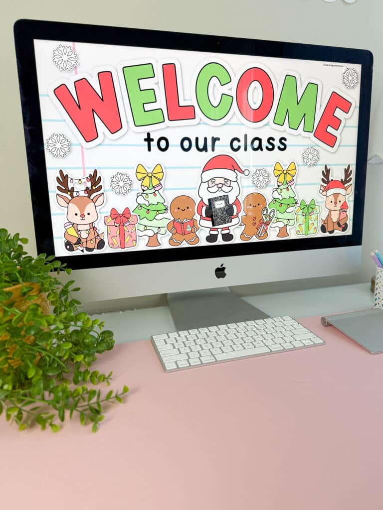 An iMac on a pink desk displays festive Christmas classroom decor with a “Welcome to our class” slide featuring reindeer, Christmas trees, gingerbread characters, gifts, and Santa holding a composition notebook. A plant sits on the left side of the desk, and a keyboard is placed in front of the computer.