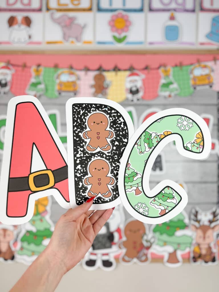 A hand holds up three festive alphabet letters—A, B, and C—decorated with Santa belts, gingerbread composition notebook patterns, and Christmas trees, displayed in front of colorful Christmas classroom decor featuring Santa, gingerbread characters, reindeer, and holiday-themed banners.