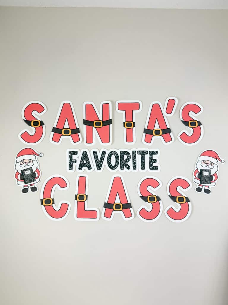 A “Santa’s Favorite Class” Christmas classroom decor display featuring large red letters with Santa belt designs and two Santa cut-outs holding composition notebooks, arranged on a light beige wall.