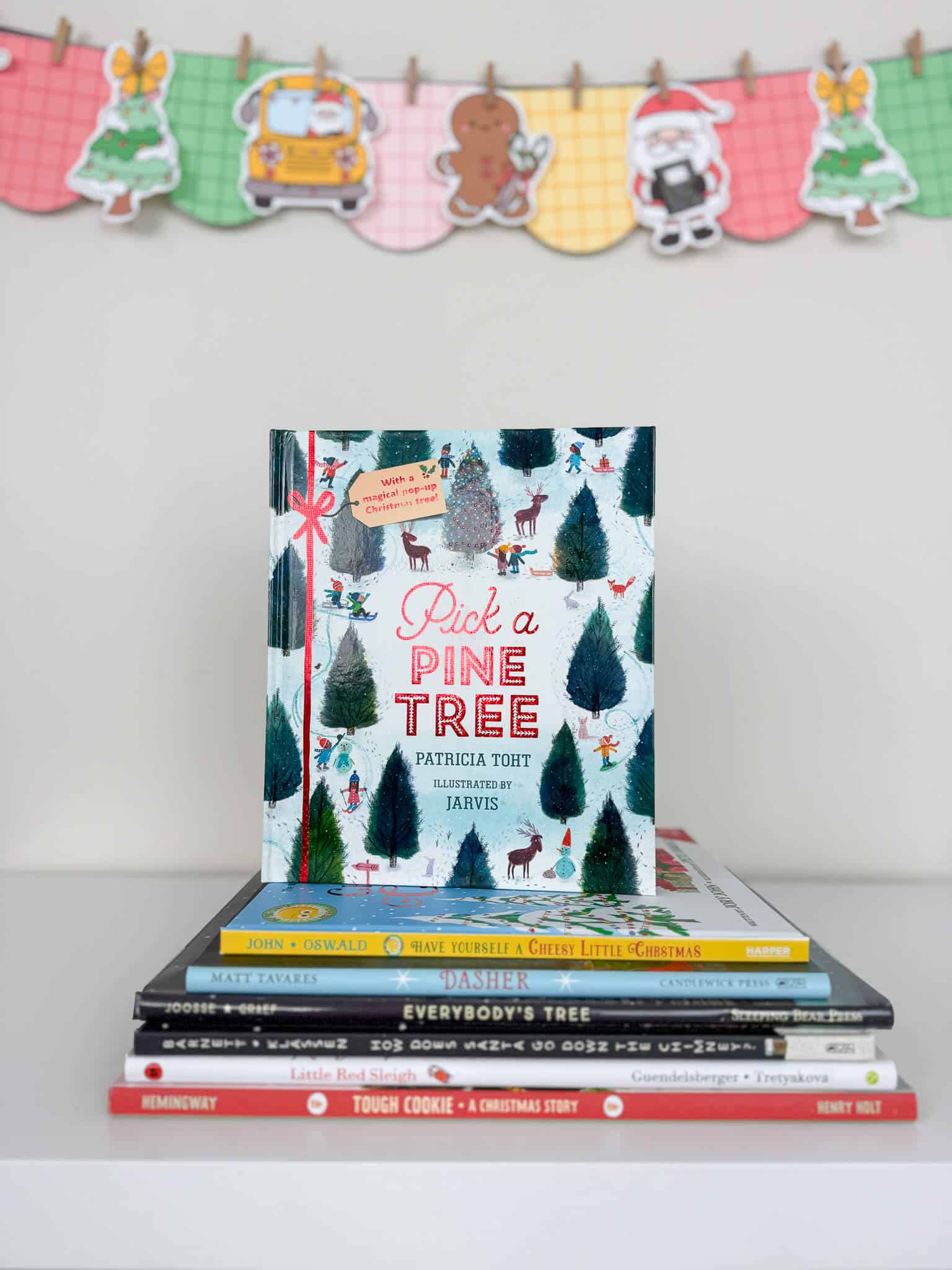 A stack of Christmas-themed children's books sits on a white shelf, with Pick a Pine Tree displayed upright on top. Behind the books is a colorful holiday banner featuring a school bus, Christmas trees, a gingerbread cookie, and Santa, all hanging on a plaid garland. Christmas Read Aloud