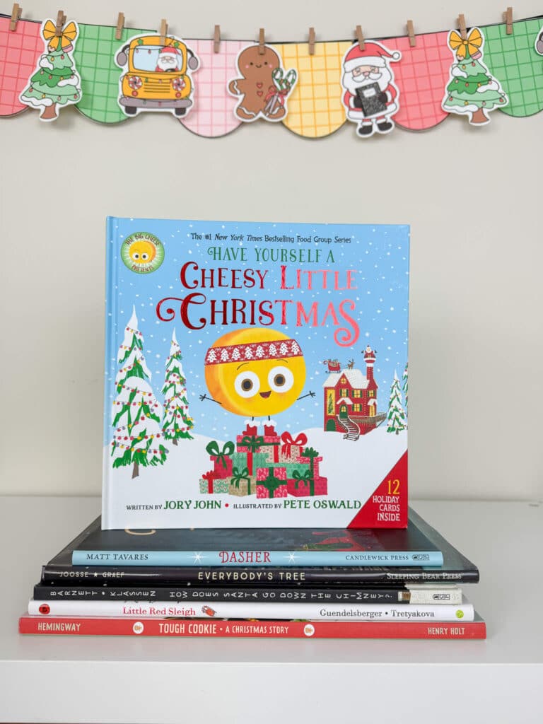 A copy of Have Yourself a Cheesy Little Christmas is displayed upright on top of a stack of Christmas-themed children's books on a white shelf. Behind the books hangs a colorful holiday banner featuring a Christmas tree, school bus, gingerbread cookie, Santa, and another Christmas tree, all on plaid-patterned pennants.