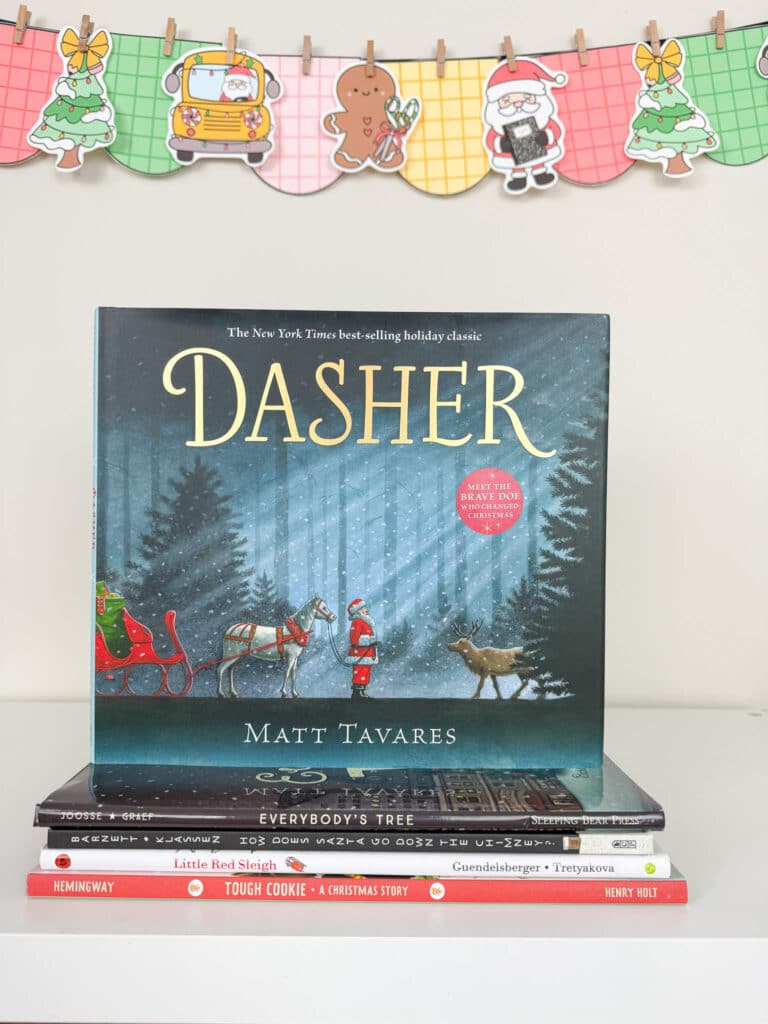 The Christmas picture book Dasher is displayed upright on a white shelf, resting on top of a stack of other holiday books. The cover shows Santa standing beside a sleigh and a reindeer in a snowy forest. Behind the books hangs a colorful holiday banner with plaid-patterned pennants featuring a Christmas tree, school bus with Santa, a gingerbread cookie, Santa holding a present, and another Christmas tree.