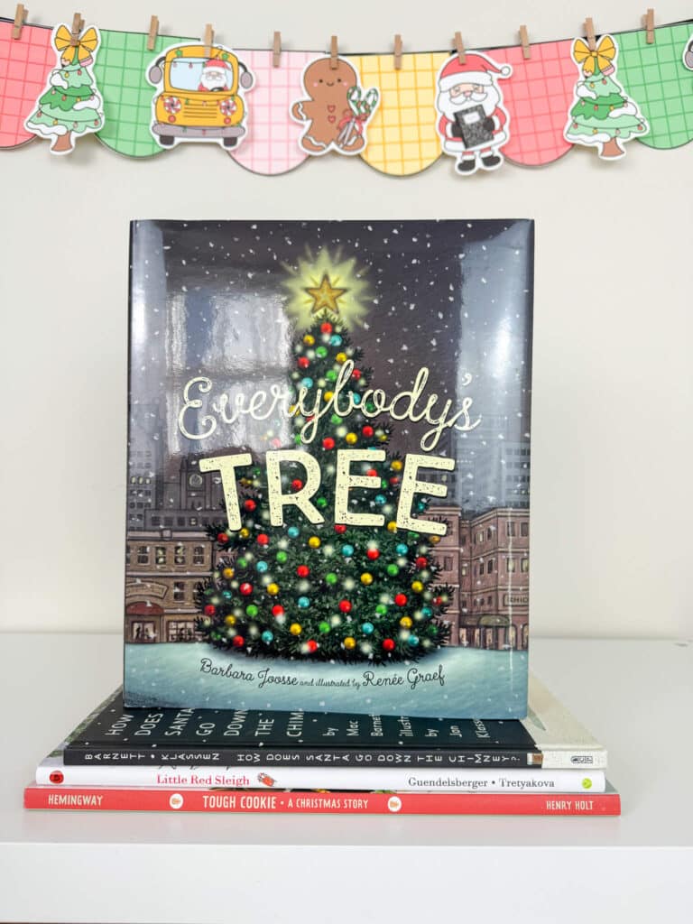 The children’s book Everybody’s Tree is displayed upright on a white shelf, showing an illustrated city Christmas tree glowing with lights and a star on top. The book rests on a small stack of holiday picture books. Behind the display hangs a colorful Christmas banner with plaid-patterned pennants featuring a tree, a school bus with Santa, a gingerbread cookie, Santa holding a present, and another Christmas tree.