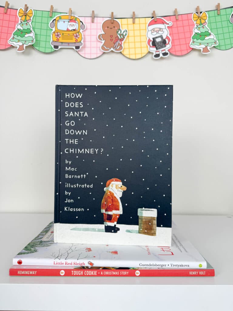 The picture book How Does Santa Go Down the Chimney? is displayed upright on a white shelf. The cover shows Santa standing in the snow, staring thoughtfully at a small chimney. The book rests on top of Little Red Sleigh and Tough Cookie. Behind the books hangs a colorful holiday banner with plaid-patterned pennants featuring a Christmas tree, a school bus with Santa, a gingerbread cookie, Santa holding a present, and another Christmas tree.
