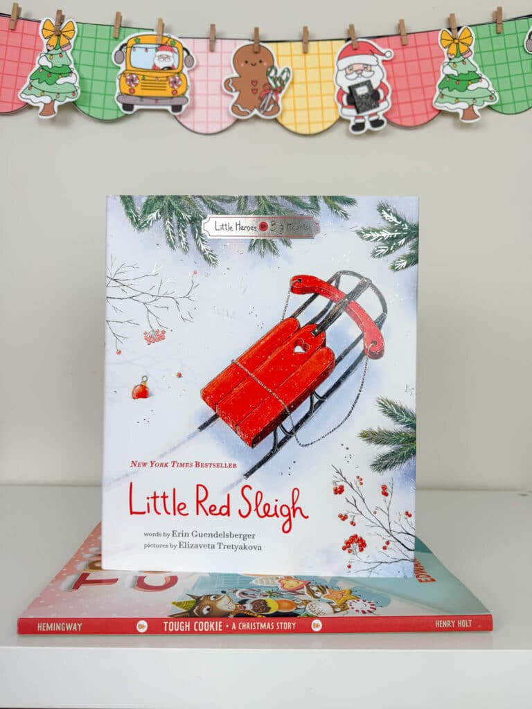 The picture book Little Red Sleigh is displayed upright on a white shelf, showing an illustration of a bright red sleigh resting in the snow beneath pine branches. The book sits on top of another holiday book, Tough Cookie. Behind the books hangs a colorful Christmas banner with plaid-patterned pennants featuring a decorated tree, a school bus with Santa, a gingerbread cookie, Santa holding a present, and another Christmas tree.