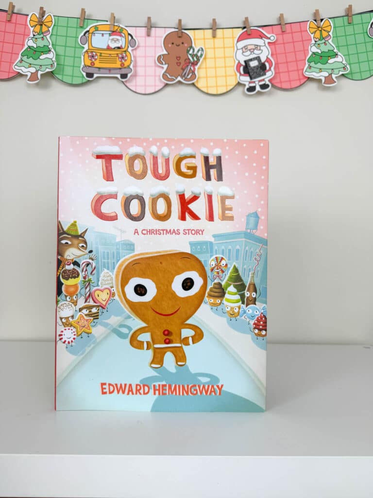 The picture book Tough Cookie: A Christmas Story is displayed upright on a white shelf. The cover features a smiling gingerbread cookie standing confidently in the snowy Land of Holiday Treats, surrounded by other illustrated cookies and sweets. Behind the book hangs a colorful holiday banner with plaid-patterned pennants showing a Christmas tree, a school bus with Santa, a gingerbread cookie, Santa holding a present, and another decorated tree.