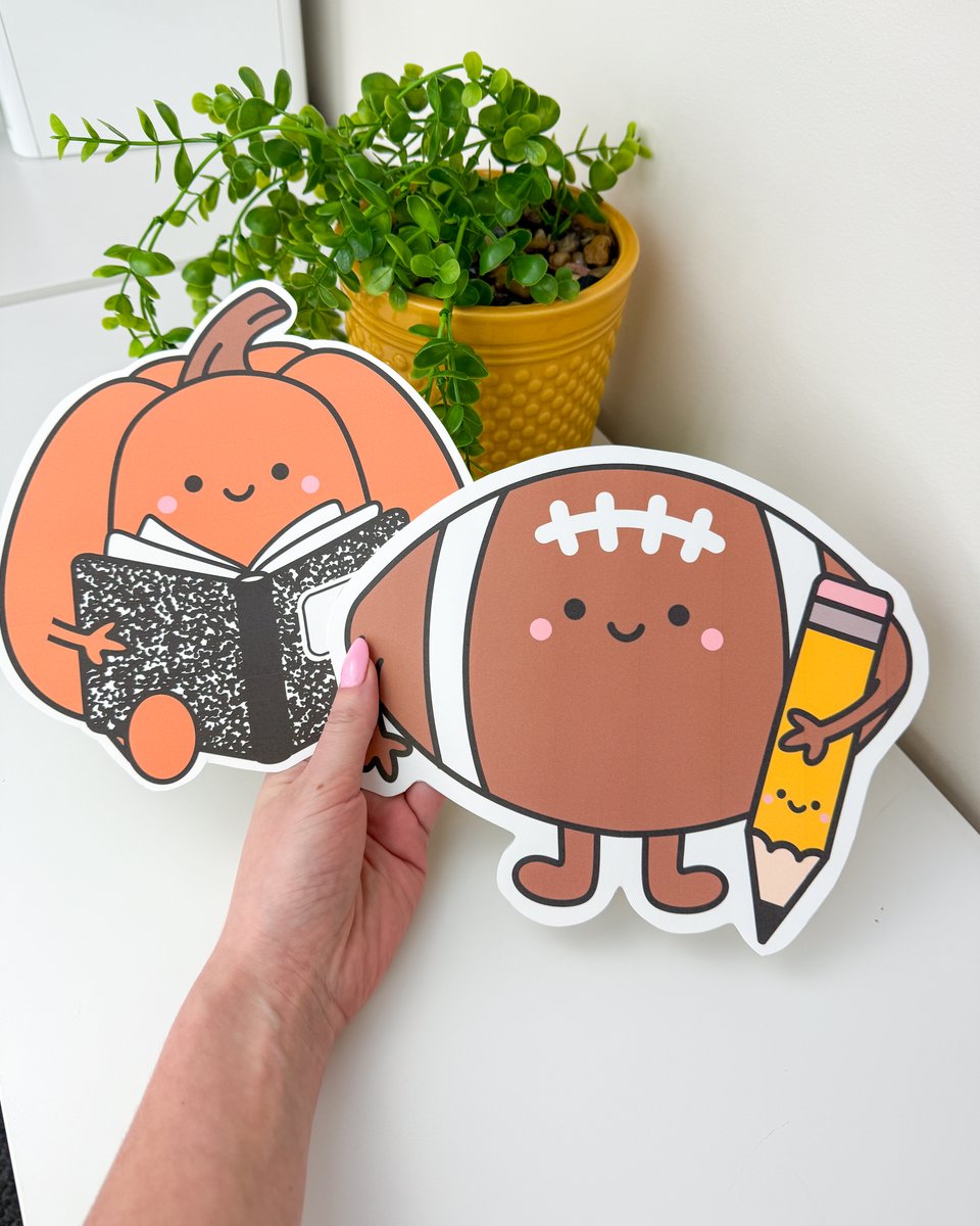 Hand holding two fall bulletin board cutouts: a pumpkin reading a composition notebook and a football holding a pencil