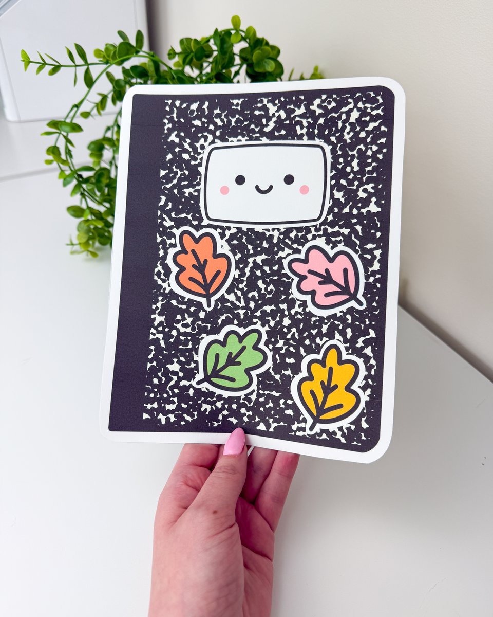 Hand holding a printed composition notebook poster with a smiling label and orange, pink, green and yellow fall leaves